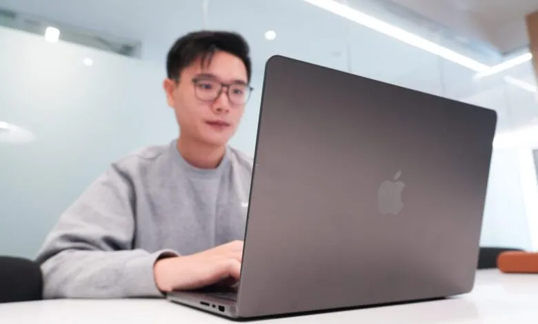 Young man wearing glasses and a grey sweatshirt works on a laptop.