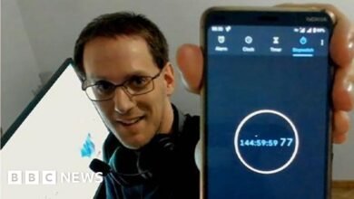 Man in glasses holds up a smartphone displaying a stopwatch at 144 hours, 59 minutes, 59 seconds.
