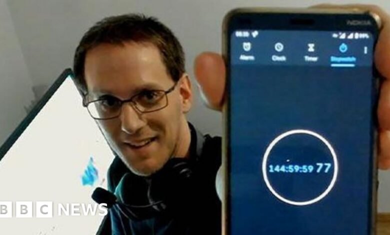 Man in glasses holds up a smartphone displaying a stopwatch at 144 hours, 59 minutes, 59 seconds.