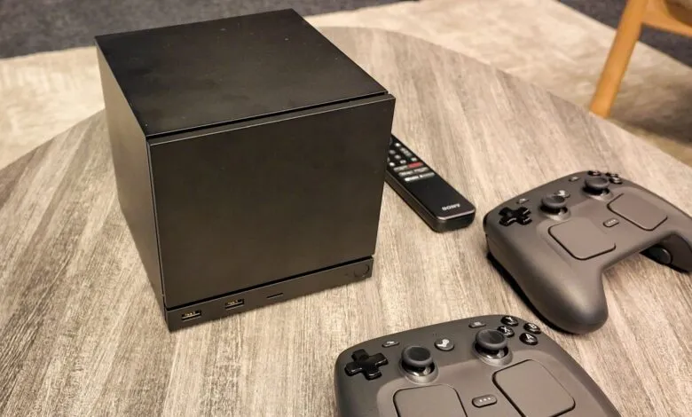 Black gaming console with two controllers and a remote on a wooden table.