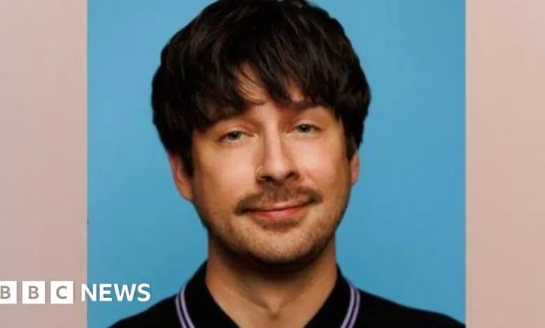 Portrait of a man with dark hair, a mustache, and a nose ring against a blue background.
