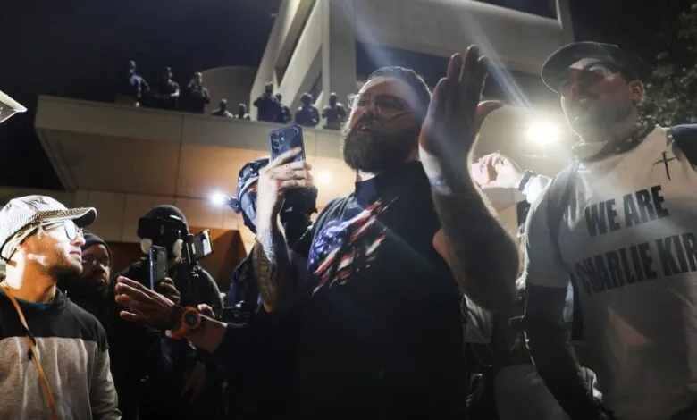 A group of people gathered at night, one man holds up a phone, another wears a shirt saying 'We Are Charlie Kirk'.