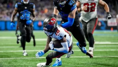 Football player in white and red uniform carrying the ball, pursued by opponents in black and blue uniforms.