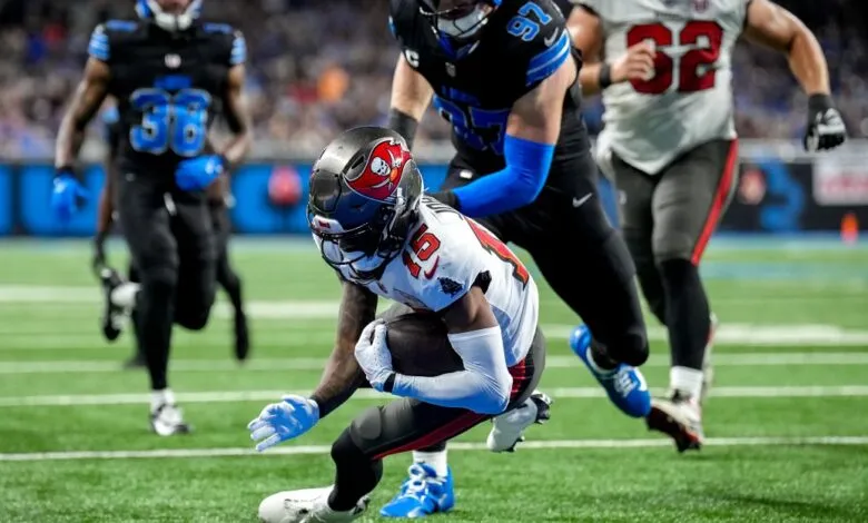 Football player in white and red uniform carrying the ball, pursued by opponents in black and blue uniforms.