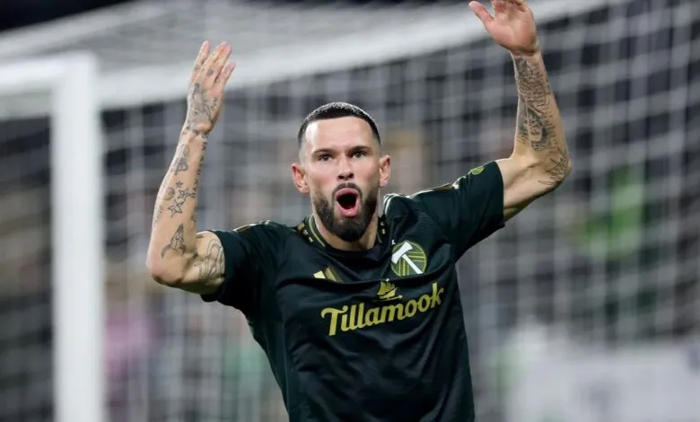 Soccer player in a dark green jersey with arms raised in celebration, showing tattoos.