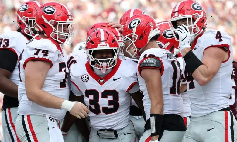 Georgia Bulldogs football players in white uniforms huddle together on the field.