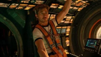 Man in a spaceship cockpit wearing a harness and looking up.