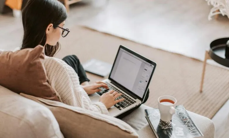 Woman wearing glasses and a sweater typing on a laptop while sitting on a couch.