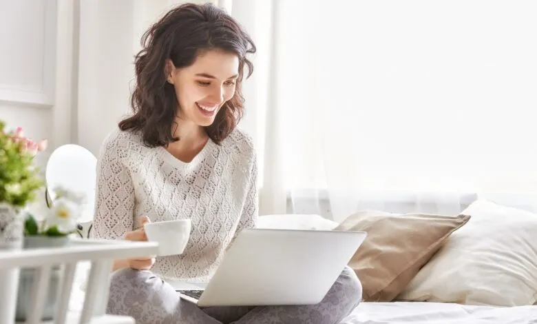 Woman smiles while working on laptop in bed with coffee.