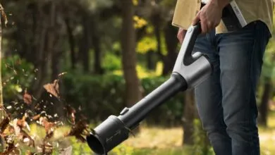 Person using a cordless leaf blower to clear fallen leaves in a wooded area.