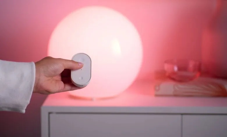 Hand holding a white smart home remote control in front of a glowing lamp.