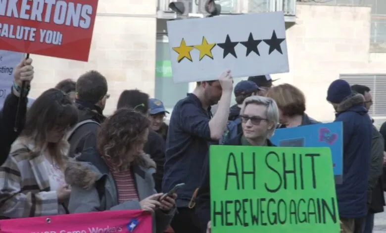 Protesters hold signs, one with two yellow stars and three black stars, another with 'AH SHIT HERE WE GO AGAIN'.