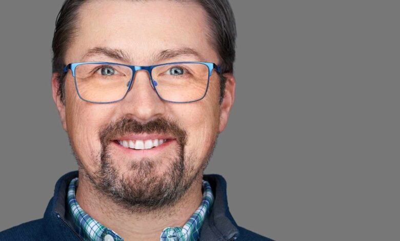 Close-up portrait of a smiling man with graying hair, beard, and blue glasses.