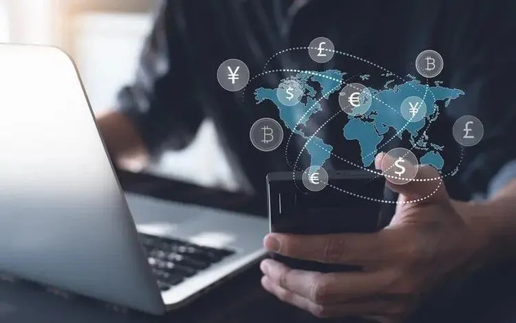 Person using a smartphone and laptop with a global currency exchange graphic overlay.