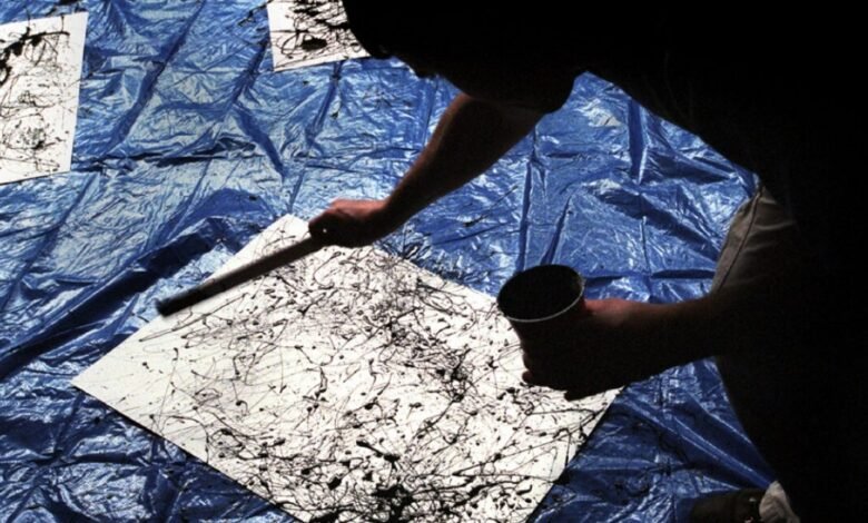 Artist dripping black paint onto canvas with a stick on a blue tarp.
