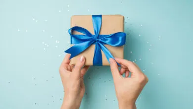 Hands untying a blue satin ribbon on a kraft paper gift box with scattered sequins.