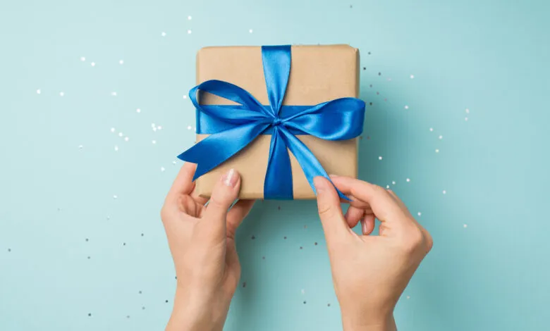 Hands untying a blue satin ribbon on a kraft paper gift box with scattered sequins.
