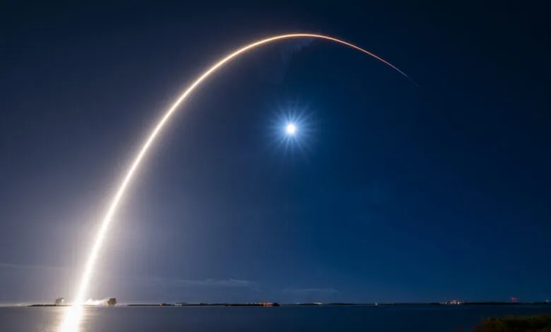 Rocket launch streak against a dark blue night sky with a bright moon.