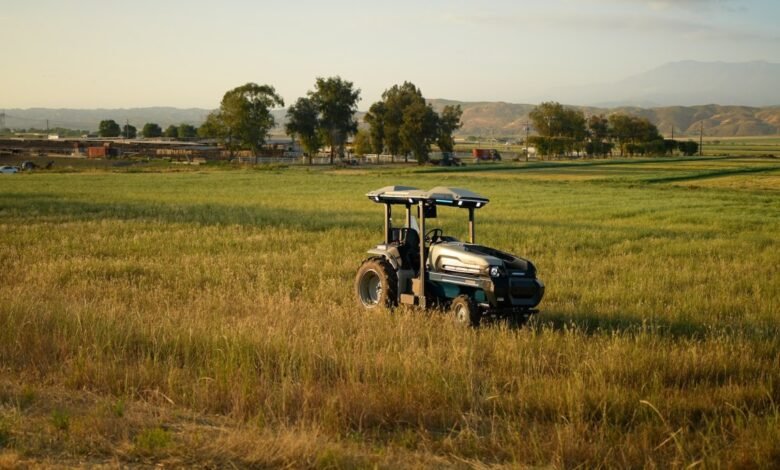 A futuristic autonomous tractor drives through a grassy field at sunset.