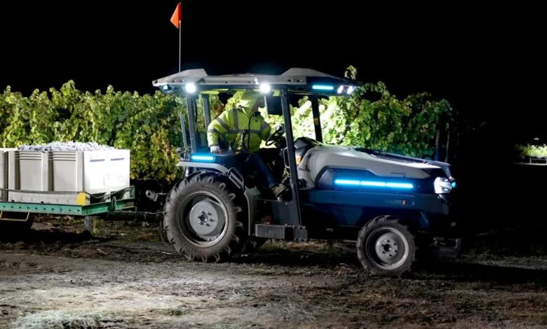 A person drives a modern tractor at night, harvesting grapes in a vineyard.