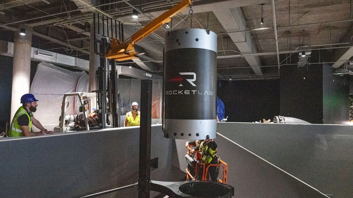 Workers in hard hats and safety vests install a large Rocket Lab component using a crane.