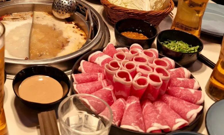 A table laden with hot pot ingredients, including thinly sliced beef, dipping sauces, and beer.
