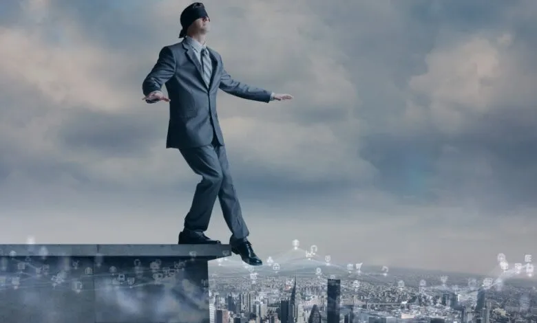 Blindfolded businessman walking on the edge of a building overlooking a city.