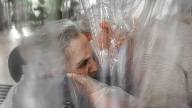 Elderly woman receives a hug through a plastic barrier.