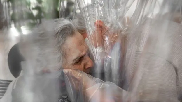 Elderly woman receives a hug through a plastic barrier.