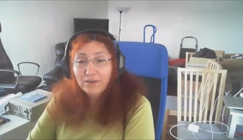 Woman with red hair and glasses wearing headphones, speaking on a video call.