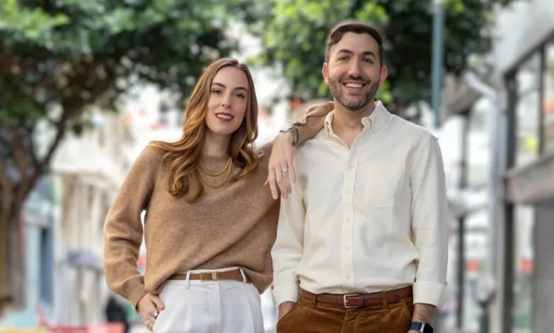 A man and a woman smiling, standing outdoors with a blurred urban background.