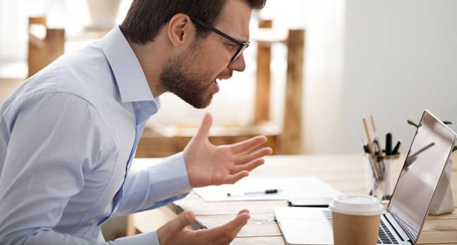 Frustrated man yelling at laptop with hands raised in anger.