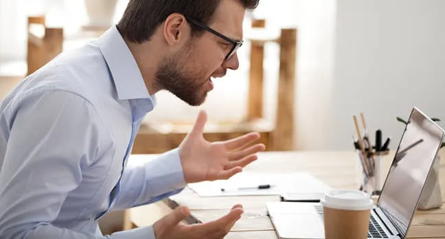 Frustrated man yelling at laptop with hands raised in anger.