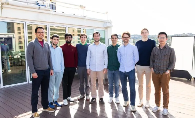 A diverse group of nine young men stand together on a wooden deck, smiling.