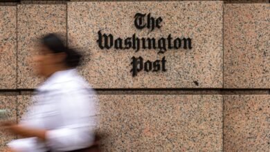 The Washington Post logo on a granite building facade with a blurred person walking by.