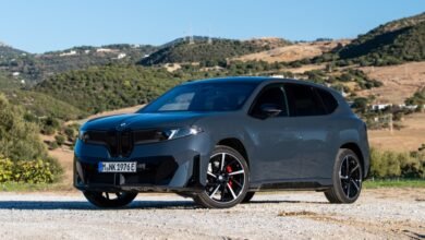 Gray BMW iX3 electric SUV parked on gravel road with hills in background.