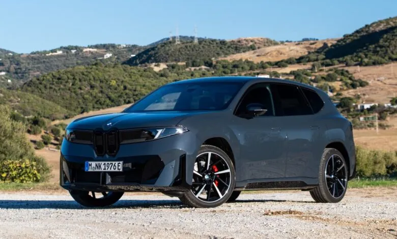 Gray BMW iX3 electric SUV parked on gravel road with hills in background.