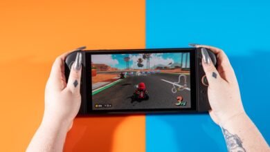 Hands holding a Nintendo Switch OLED playing Mario Kart on orange and blue background.