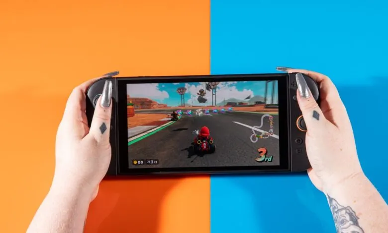 Hands holding a Nintendo Switch OLED playing Mario Kart on orange and blue background.