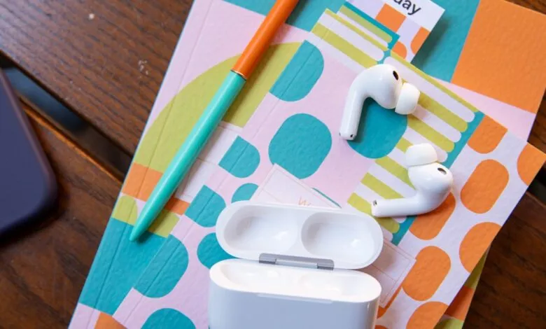 AirPods Pro with open case, pen, phone, and colorful notebooks on a wood surface.