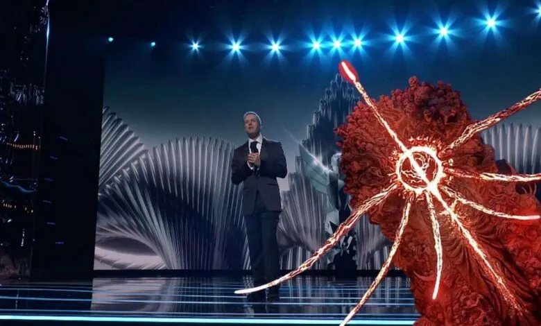 Man in suit stands on stage with abstract art and bright lights.