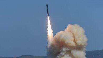 Missile launches with bright flames and smoke against a clear blue sky.