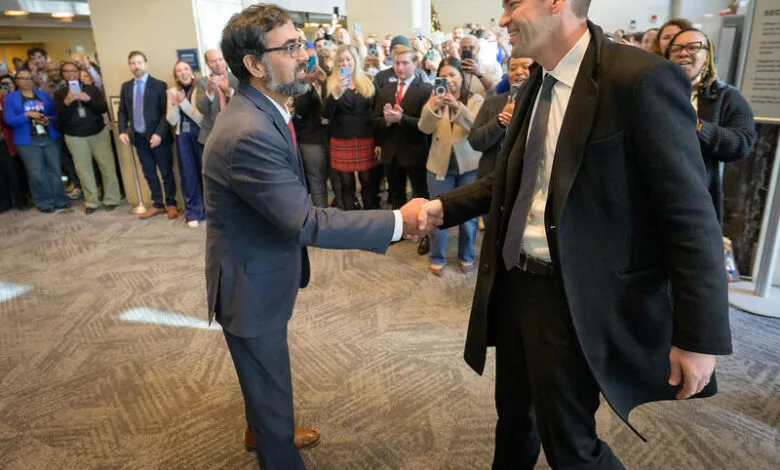 Two men shake hands in a crowded room, surrounded by onlookers taking photos.