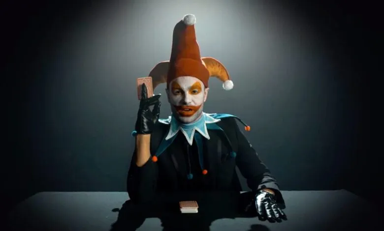 Man in jester costume holding playing card, dark background.