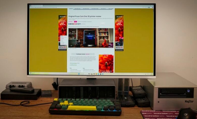 Desk setup with monitor displaying 3D printer review and various tech gadgets.