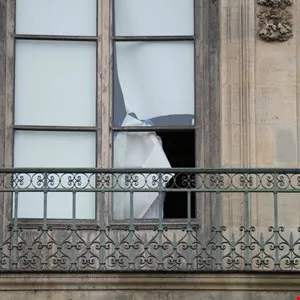 Balcony with ornate railing and a window with broken glass and white fabric.