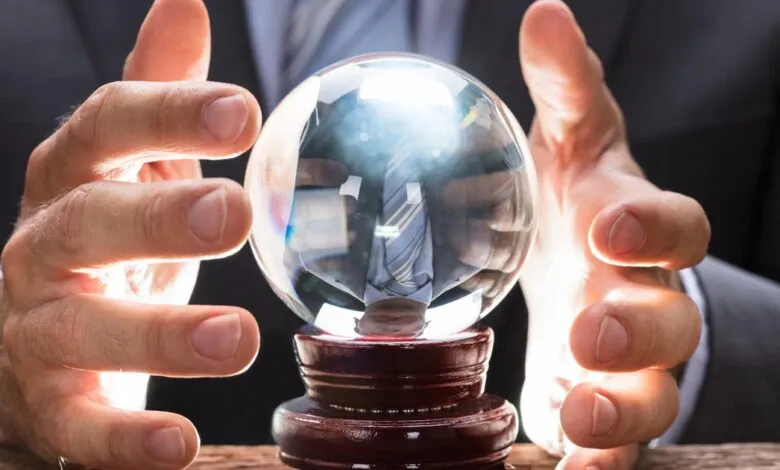 Hands surround a crystal ball on a wooden stand, reflecting a suited figure.