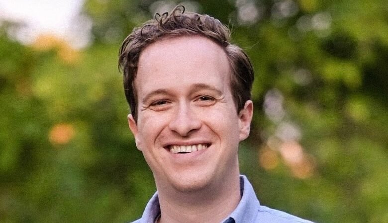 Smiling man with brown hair and blue shirt outdoors.