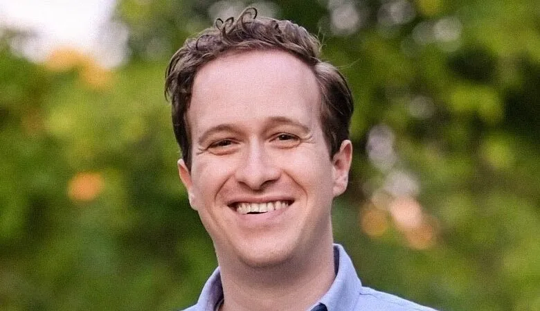 Smiling man with brown hair and blue shirt outdoors.