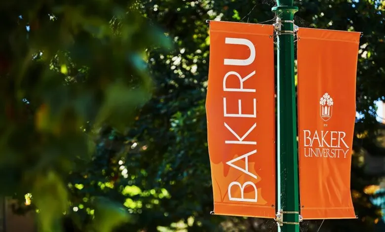 Two orange banners for Baker University hang from a green pole outdoors.